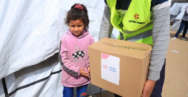 Giving Hands worker holding an aid box while a child stands beside them near a tent