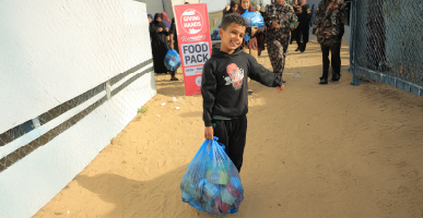 Boy carrying a blue food pack bag along a sandy corridor at a distribution point