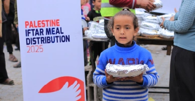 Child holding a foil-wrapped iftar meal in front of a Palestine Iftar Meal Distribution 2025 banner