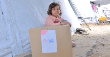 Young child sitting beside a large Giving Hands aid box inside a sandy tented area