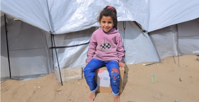 Young child sitting on a Giving Hands aid box inside a tent on sandy ground