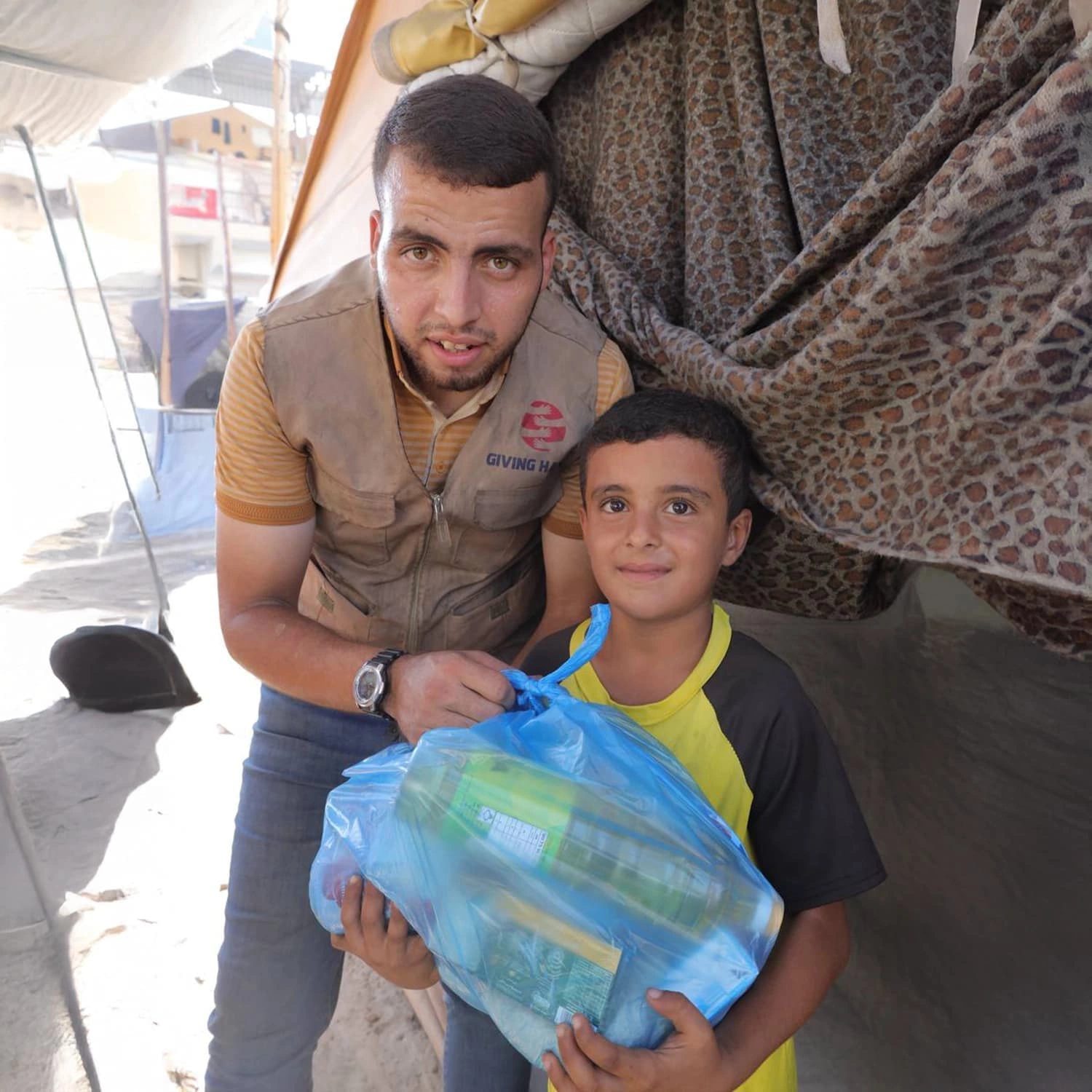 Giving Hands worker standing beside a child holding a blue relief parcel outside a tent shelter