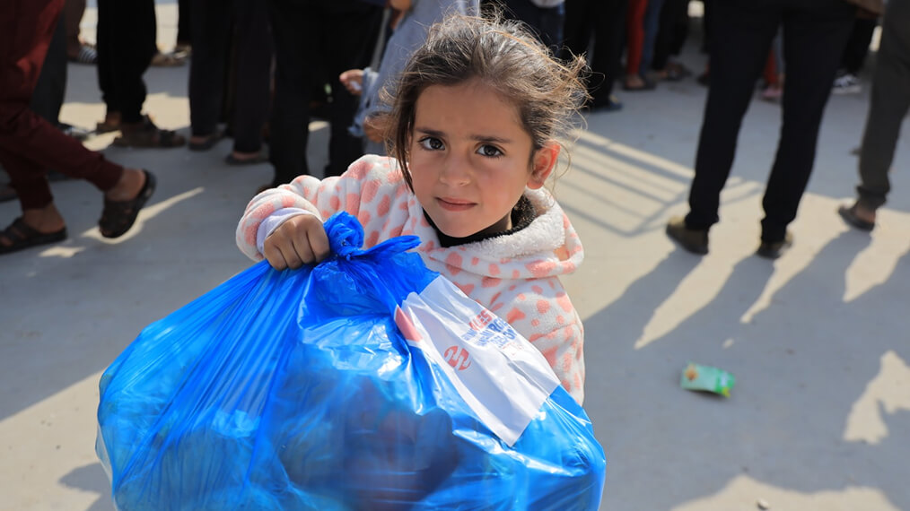 Child carrying a large blue aid bag while people stand in line in the background