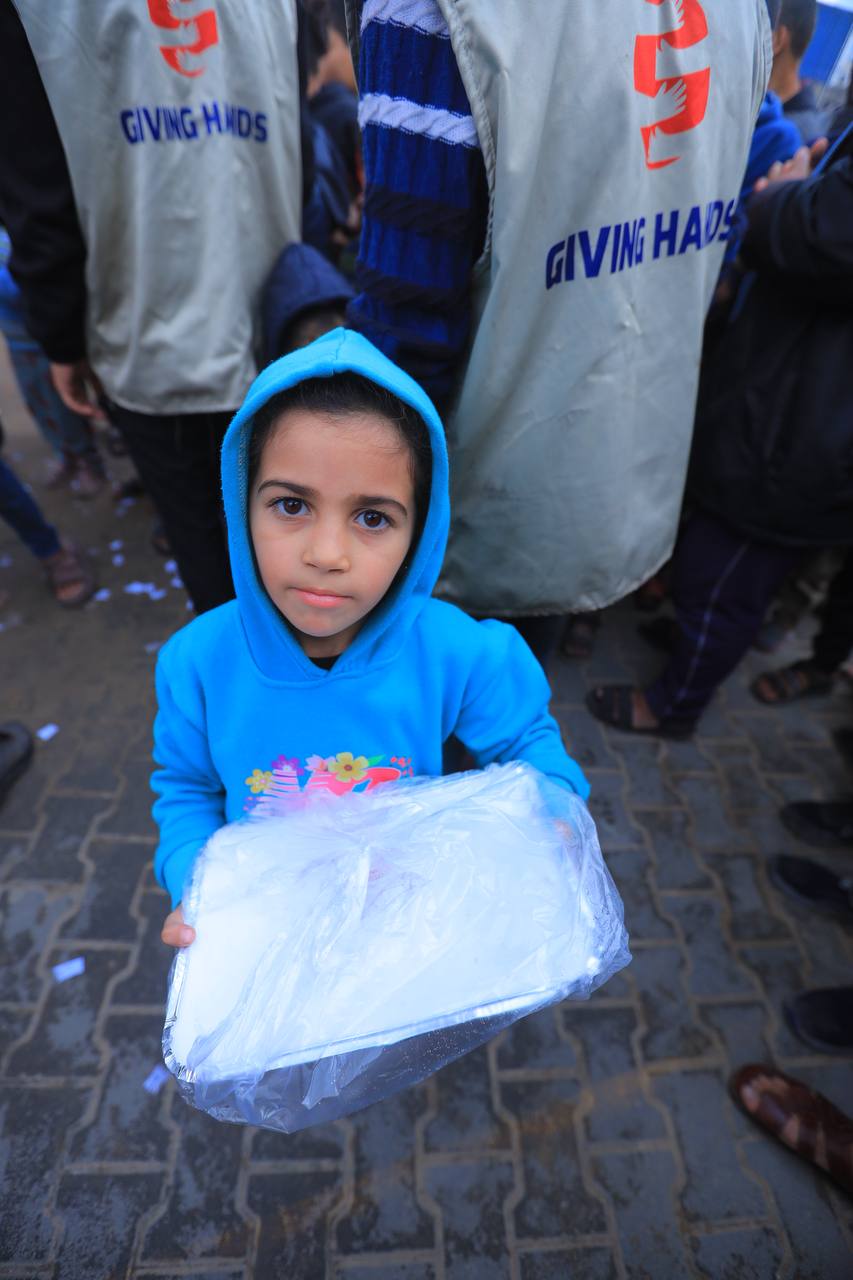 Child holding a wrapped food tray during a Giving Hands distribution on a paved street