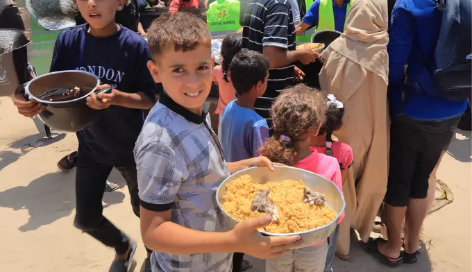 Children standing in line outdoors holding large trays of cooked rice during a Ramadan food distribution organized by Giving Hands